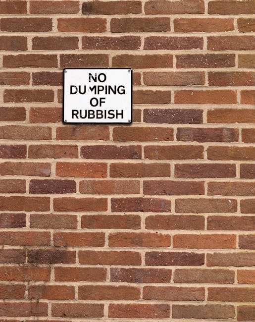 A rectangular white metal sign with black text reading 'NO DUMPING OF RUBBISH' is mounted on a red brick wall. The bricks are arranged in a traditional running bond pattern, with varying shades of reddish-brown, some darker and some lighter, creating a textured surface. The sign is positioned slightly above the middle of the image, with the brick wall extending evenly in all directions. The wall appears to be part of a building exterior or boundary, with no additional objects visible in the scene. The lighting is natural, casting soft shadows on the bricks. This image likely relates to private waste management practices or regulations concerning rubbish disposal in a residential or commercial area, as indicated by the presence of the sign emphasizing rubbish control, which is relevant to services like those provided by Rubbish Clearance Clapham.