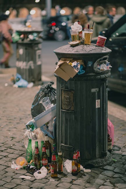 A tall, cylindrical black waste bin with a domed, textured surface is situated on a cobblestone pavement in an urban setting. The bin's lid is covered with various discarded items, including clear plastic cups filled with liquid, some containing ice and straws, and a small cardboard box. Surrounding the base of the bin are multiple empty glass bottles, including green, brown, and clear varieties, along with crumpled paper, plastic wrappers, and other waste. A black garbage bag is tied and placed next to the bin, with additional waste scattered on the ground. In the background, a blurred city street scene is visible, featuring parked cars and people walking, with a large municipal trash container or bin nearby. The environment suggests an outdoor public area, possibly in the vicinity of a café or busy pedestrian zone, highlighting issues around informal rubbish disposal and the need for regular waste collection.