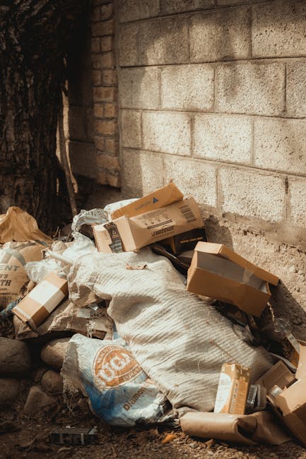 A pile of mixed waste materials resting against a weathered brick wall outdoors. The collection includes various cardboard boxes, some flattened and others partially collapsed, with visible tape and printed labels. There are also several large plastic bags, one with an orange and white logo, filled with rubbish and tied at the top. The ground beneath the debris consists of small rocks and dirt, with a portion of a wooden tree trunk visible on the left side of the image. The surrounding environment appears to be an exterior space, possibly an alley or side yard, with natural lighting casting soft shadows. This scene relates to private waste disposal and rubbish removal services, highlighting the type of general waste that might be collected by companies like Rubbish Clearance Clapham during on-site clearance or alternative rubbish collection efforts outside of municipal waste services.