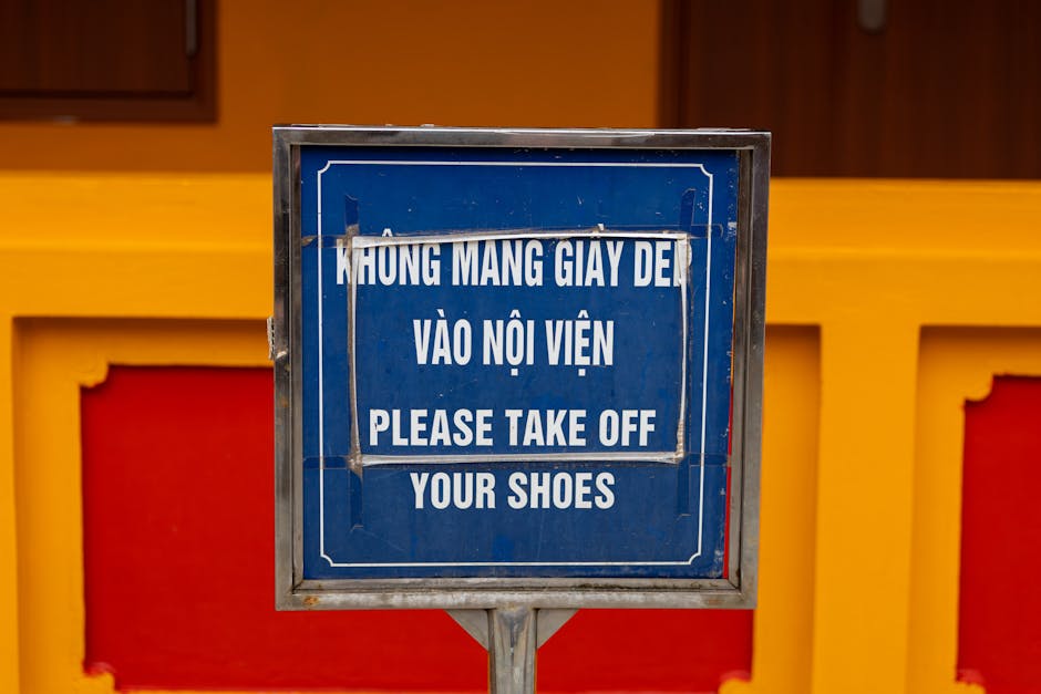 A square blue sign with a white border and white text in both Vietnamese and English, positioned on a metal frame in front of a vibrant yellow and red wall. The sign reads 'HƯNG MANG GIÀY DẸ VÀO NỘI VIỆN PLEASE TAKE OFF YOUR SHOES.' The sign appears slightly weathered with minor creases and scratches. The background features portions of a decorative wall with rectangular panels, some painted red and others yellow, suggesting a cultural or religious setting. The lighting is natural and even, indicating the photo was taken indoors or in a shaded outdoor area. This scene may relate to the need for proper disposal of footwear or the etiquette of removing shoes before entering certain premises, which aligns with the themes of private waste handling or on-site clearance services provided by Rubbish Clearance Clapham.