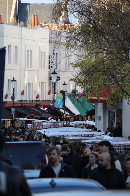 A busy outdoor marketplace scene with numerous market stalls covered with white and green canopies, closely packed together along a pedestrian pathway. The foreground shows a crowd of people browsing and walking, with some individuals partially blurred to indicate movement. To the left, there are several black street lamps mounted on the white building facade, which features rectangular windows and a vertical sign with the word 'BAR'. The background includes a multi-storey building with a plain white exterior, visible chimney stacks on the rooftop, and some trees with autumn foliage partially obscuring the view. The scene is lit by natural daylight, casting soft shadows, and suggests a lively urban setting fitting for an event like a market or fair, with the environment resembling an outdoor gathering area where waste management or rubbish removal services, such as those provided by Rubbish Clearance Clapham, might be required to maintain cleanliness amid the crowds and activity.